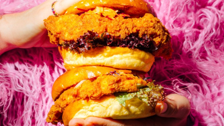 TDR-WOMO, © wenigistmehr Two large, deep-fried chicken burgers are held by hands in front of a pink, fluffy background.