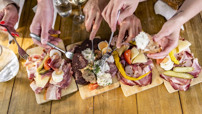 Heurigen snack, © Weinviertel Tourismus GmbH / Herbst Several hands reach for platters of sausage and cheese on a wooden table.