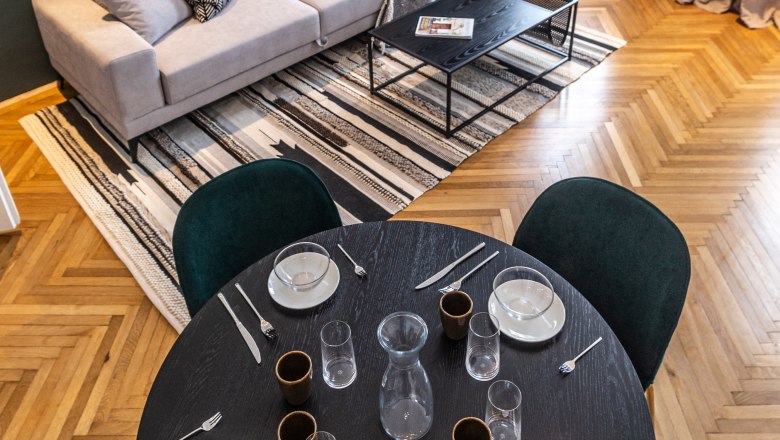 Apartment, © Astrid Bartl Modern living room with sofa, coffee table, carpet and dining table with place settings.