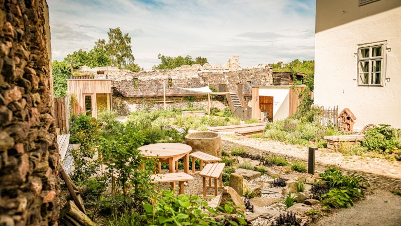Spend the night in the city loft, © SONNENTOR A garden with wooden tables and benches, surrounded by old stone walls and plants, under a blue sky.