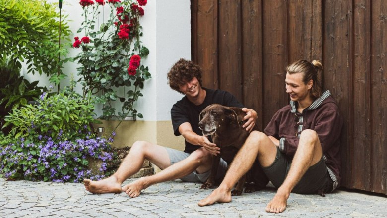 Florian and Lorenz Widerna, © Elke Wolfbeisser Two men sit barefoot on a cobbled floor next to a brown dog. Red roses and a wooden wall can be seen in the background.