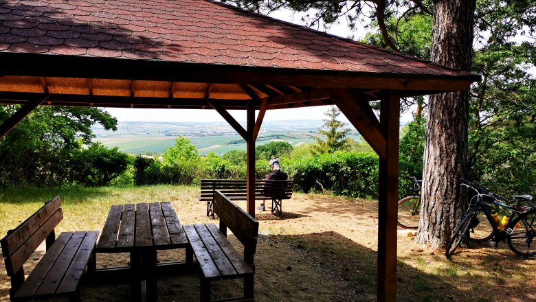 On some days you can even see as far as the Ötscher, © Weinstraße Weinviertel Wooden pavilion with table and benches, view of vineyards, bicycle on a tree.