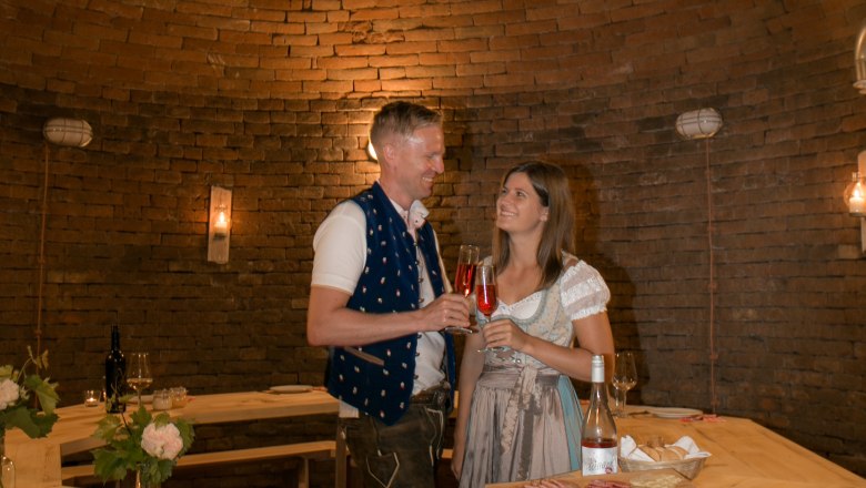Winery Küssler, © Winzerhof Küssler A couple in traditional dress toast with drinks in a rustic, vaulted room.