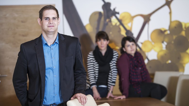 The Blaha family, © Alfred Kusdat A man in a blue shirt and black jacket stands in front of a table, with two women sitting in the background.