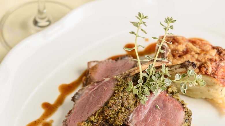 Dining view Hotel-Restaurant Zur Post, © BOW Rita Newman A plate with herb-crusted lamb chops, vegetable garnish and sauce, with a glass of red wine next to it.