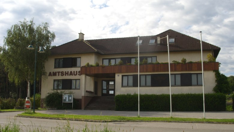 Natschbach-Loipersbach office building, © Gemeinde Natschbach-Loipersbach The Natschbach-Loipersbach office building with brown roof and balcony, surrounded by trees and flagpoles.