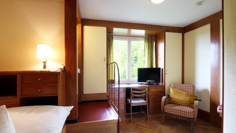 Double room, © Christian Redtenbacher A cozy hotel room with wooden furniture, a TV, an armchair and a window with a view of the greenery.