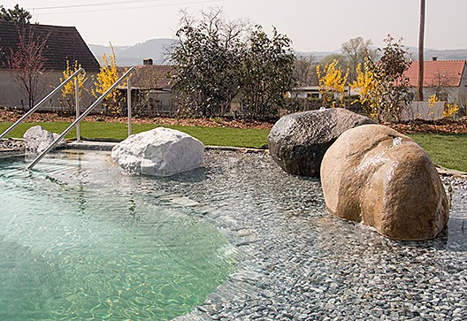 Swimming pool, © Weinresidenz Sonnleitner A swimming pool with large stones and a metal ladder, surrounded by plants and a garden.