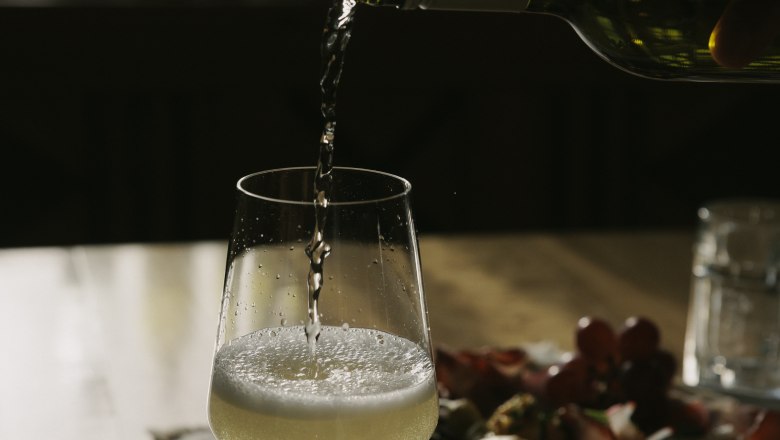 Pouring wine, © Weinviertel Tourismus / Sophie Menegaldo A glass of white wine is poured on a table, with a plate of snacks in the background.