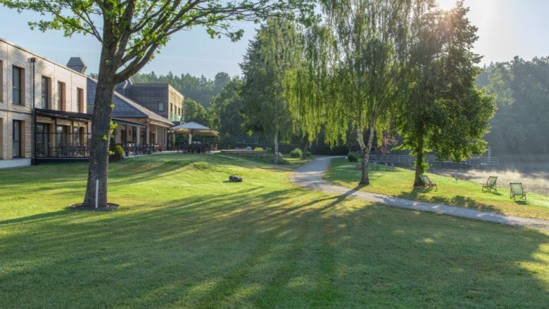 Hotel-Restaurant Hausschachen, © Hotel-Restaurant Hausschachen Green meadow with trees, buildings and deckchairs on the lakeshore in the sunlight.