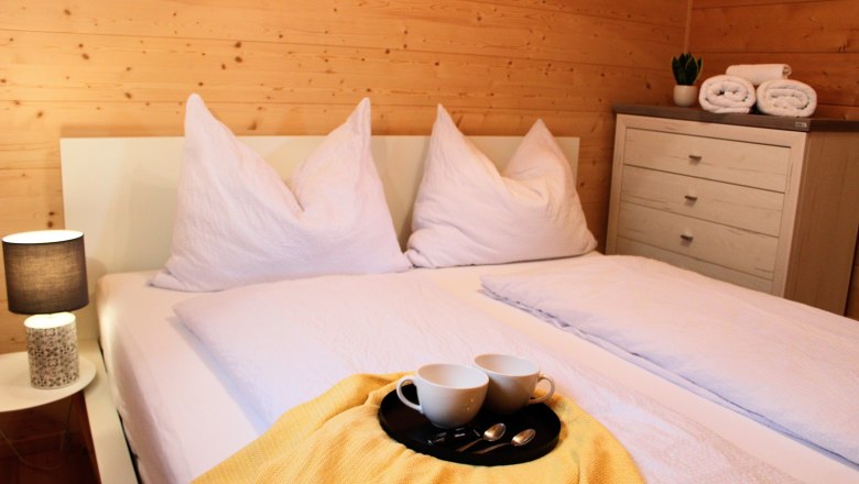 Bedroom, © Fam. Auer Cozy bedroom with double bed, white pillows and blankets, yellow blanket, tray with cups, bedside lamp and chest of drawers with towels.