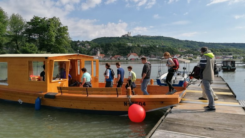 Access to the Zille, © Schiffsführerschule Ing. Karl Hell People board a wooden boat on a jetty, in the background a hilly landscape with buildings.