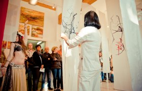 Atelier Buchner, © Christine Buchner, Foto Birgit Muehleder A woman dressed in white looks at drawings on hanging fabric panels in a gallery with spectators and paintings on the wall.