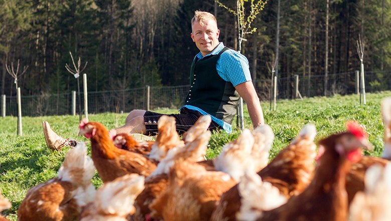 Christoph Holzbauer, © Sooo gut schmeckt die Bucklige Welt/ Viktoria Kornfeld A man in traditional costume sits in a green meadow with chickens in the foreground.