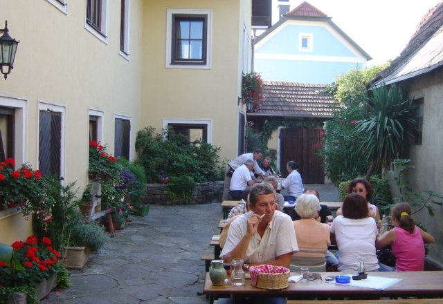 Winery, © Fam. Supperer People sit at wooden tables in a courtyard with yellow buildings and plants.