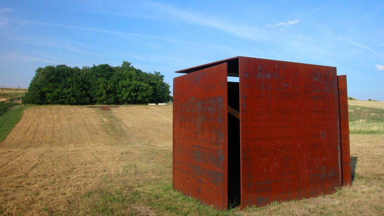 Wind cube house, © Stadtgemeinde Mistelbach A rusty metal sculpture stands in a field in front of a forest under a blue sky.