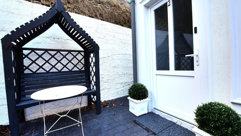 Top 2 Outdoor area - entrance, © Daniela Sinadinoska Entrance area with white door, black bench with canopy and small table.