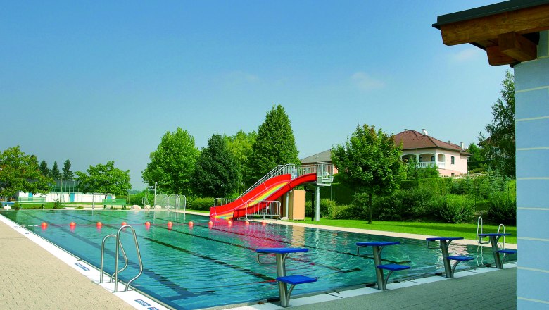 Outdoor pool Unterstinkenbrunn, © Gemeinde Unterstinkenbrunn An outdoor pool with a swimming pool, a red slide and starting blocks. Trees and a house can be seen in the background.