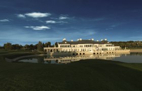 Clubhouse, © FONTANA An elegant clubhouse is reflected in a tranquil lake, surrounded by green lawns and trees under a clear blue sky.
