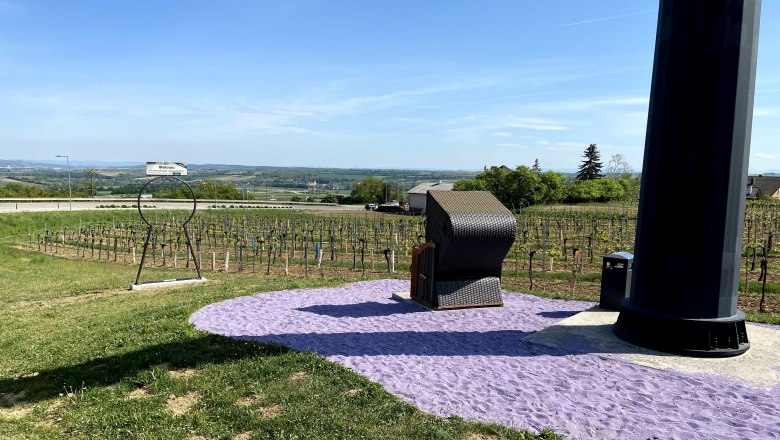 Relaxing in a beach chair on the purple sand of Maissau, © Weinstraße Weinviertel A beach chair stands on purple sand next to a large black pillar, surrounded by vineyards and a vast landscape.