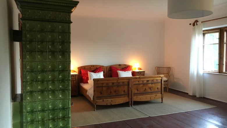 Double room, © Riegelhof A cozy double room with wooden furniture, a green tiled stove and a large window with white curtains.