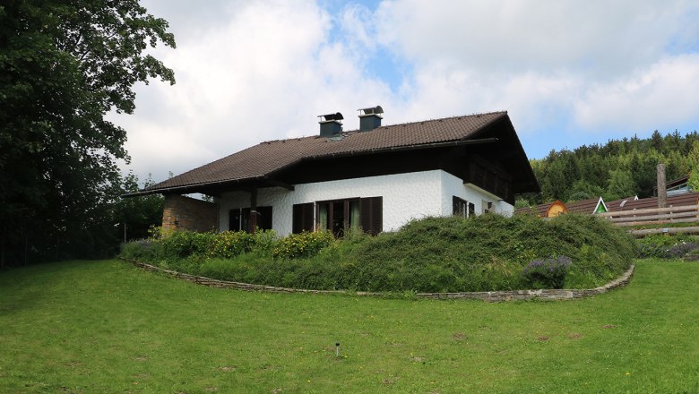 Home garden, © Pamela Hniliczka A white house with a brown roof in a green garden with trees and flowers.