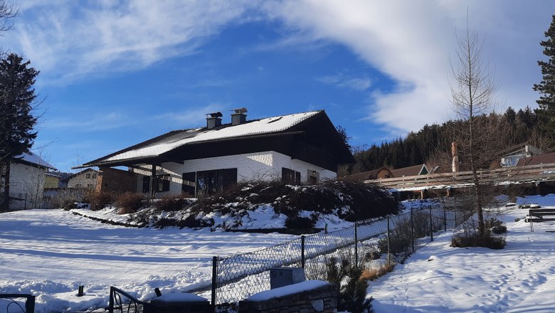 Belle Air Lodge in winter, © Christoph Gierlinger A snow-covered house with a dark roof and chimneys, surrounded by trees and a blue sky.