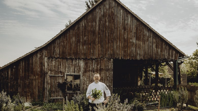 Inn with Weinviertel cooking courses, © Niederösterreich Werbung/Sophie Menegaldo A cook stands in front of an old wooden hut with a bunch of herbs in his hand.