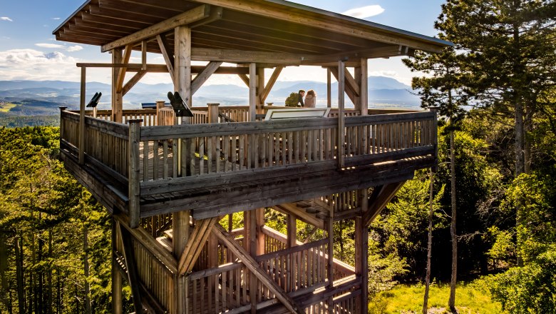 The observation tower in Lanzenkirchen/Wiesen, © Wiener Alpen/Fülöp, Kremsl A drone shot from the side of the observation tower, on which two people are standing. Green forest and mountains in the background