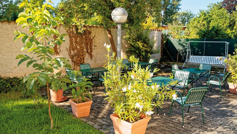 Garden, © Christoph Sammer A sunny garden with green chairs, tables and plants in pots on a paved terrace.