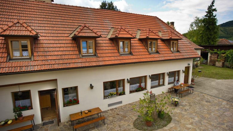 Inner courtyard, © Dinstlhof Georg Edlinger A traditional building with a red tiled roof and skylights, surrounded by a paved courtyard with tables and plants.