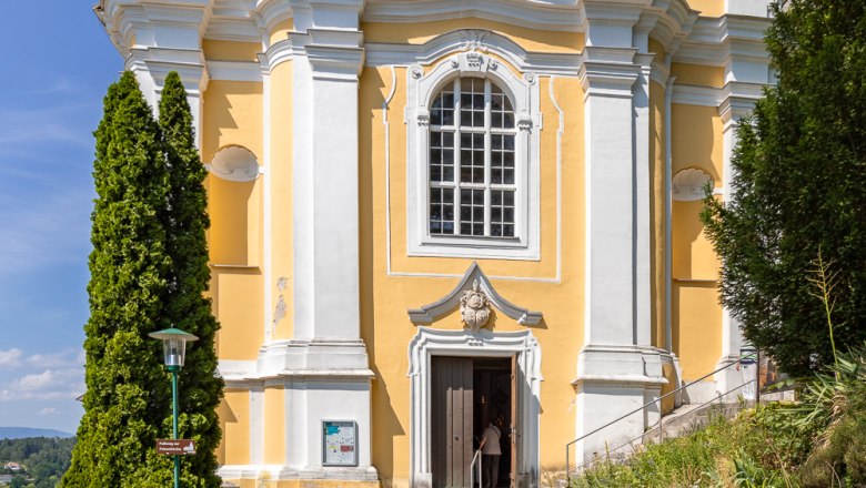 Mountain church Pitten, © Wiener Alpen, Christian Kremsl Yellow baroque church with white columns and a statue above the entrance, surrounded by trees and blue sky.