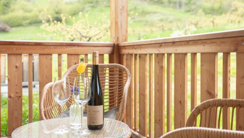 View from the balcony, © Weingut Rixinger A balcony with wooden furniture, a bottle of wine and glasses on a table. Green landscape in the background.