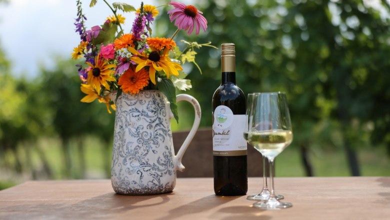 Idyllic wine tavern, © Manuela Winkler Wine bottle and glass on table with bouquet of flowers in vase, grapevines in background.