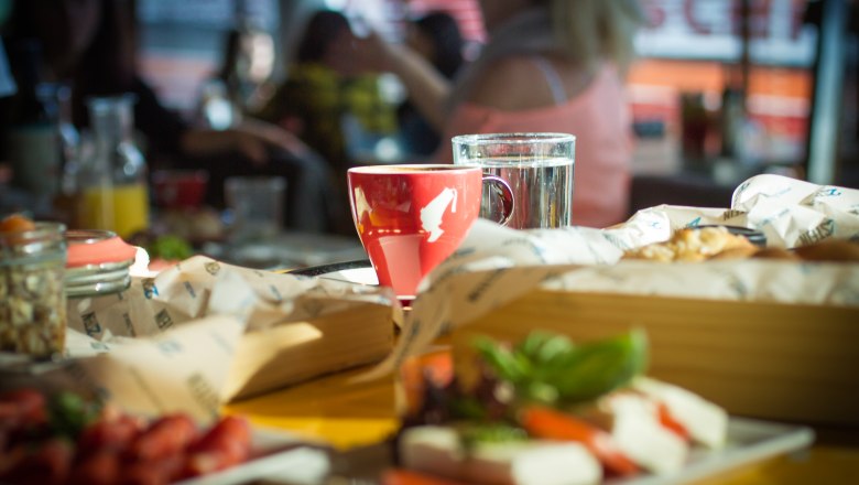 2Stein Bar-Café-Restaurant, © 2stein_Kastner new media A red coffee mug and a glass of water on a table in a café, surrounded by food and drinks.