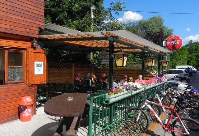 XLarge snack bar Kritzendorf, © Brigitta Moretti Outdoor area of a snack bar with wooden walls, tables and bicycles. People sitting under a covered area. Coca-Cola sign visible.