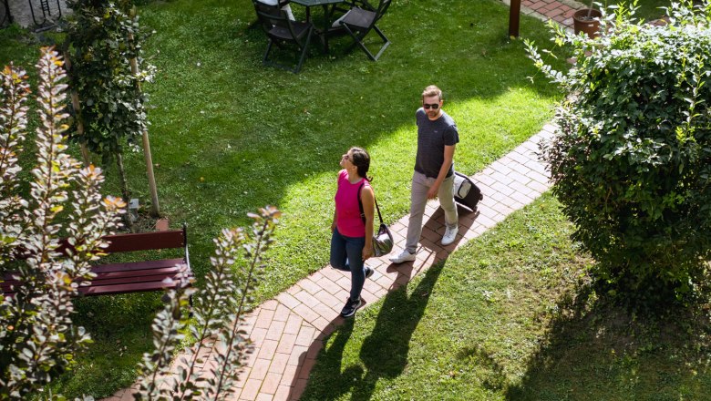 Arrival at the inn "Zum goldenen Anker", © Martina Siebenhandl Two people are walking on a paved path through a green garden with a table and chairs.
