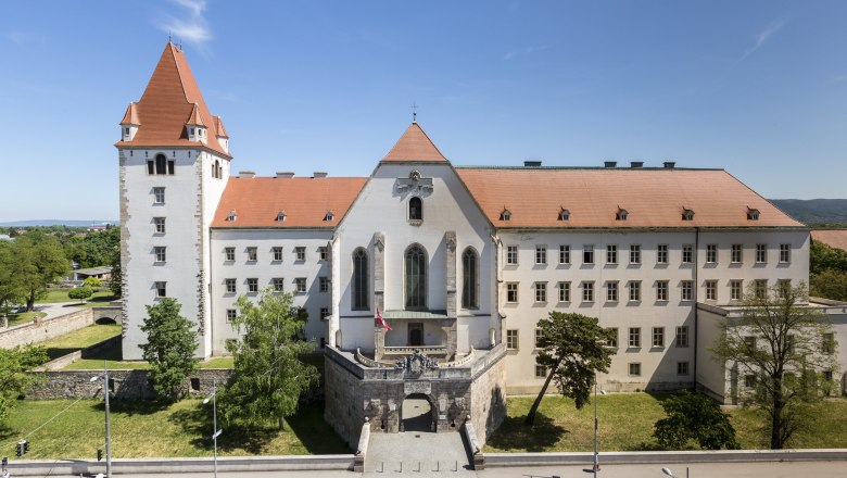 Military Academy West Gate, © Wiener Alpen, Foto: Franz Zwickl Historic building with red roof and tower, surrounded by trees and a road in the foreground.