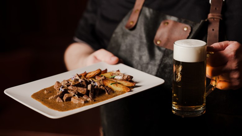 Fried liver with roast potatoes, © Niederösterreich Werbung/Michael Reidinger Person holding plate with meat dish and beer mug.