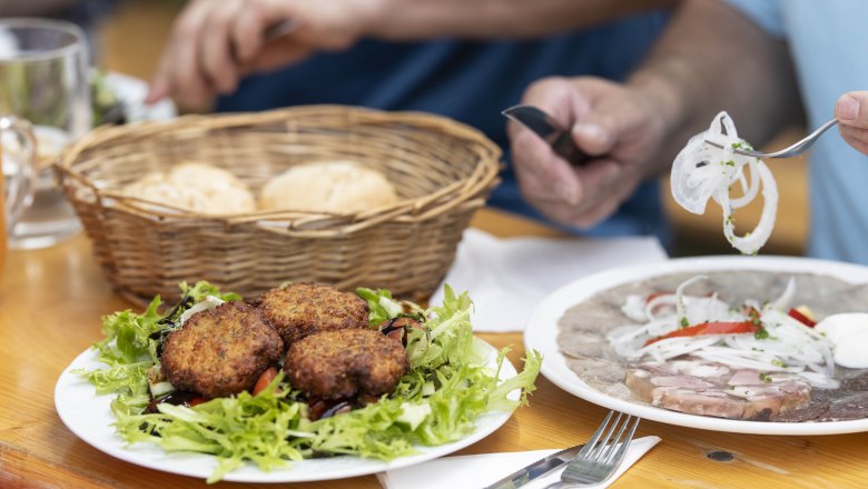 Enjoy a wine tavern snack at Genuss Heuriger Simon., © Thomas Gobauer-Photography A table with a plate full of meatballs on salad, a basket of pastries and a plate of pressed sausage and onions.