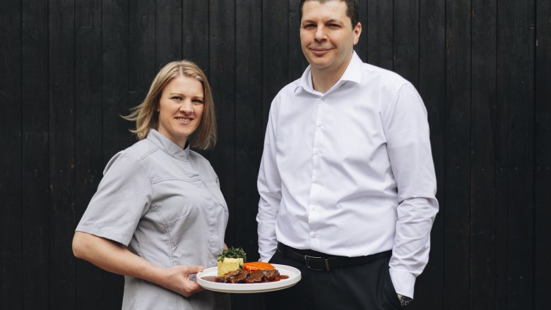 Host family Renner, © Niederösterreich Werbung/David Schreiber Two people, a woman in cooking clothes with a plate, in front of a dark background.