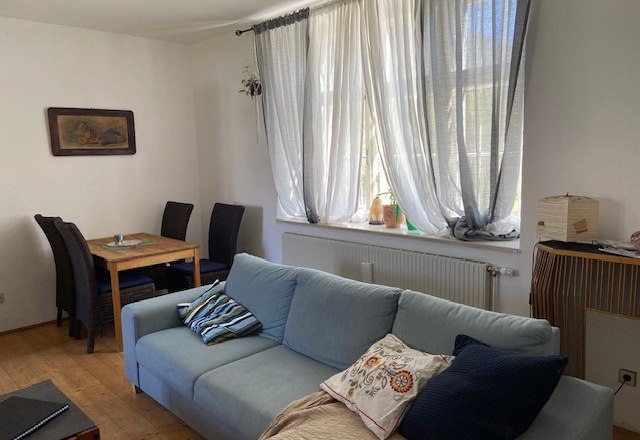 Living room, © Wiener Alpen A living room with a blue sofa, a wooden table with chairs and a window with white curtains.