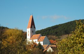 Nußdorf parish church, © Blesl Nußdorf parish church with red roof and clock tower, surrounded by trees and hills.