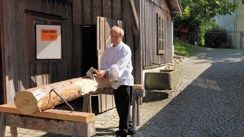 Living craftsmanship, © FVV A man is working on a tree trunk with an axe in front of a wooden house.