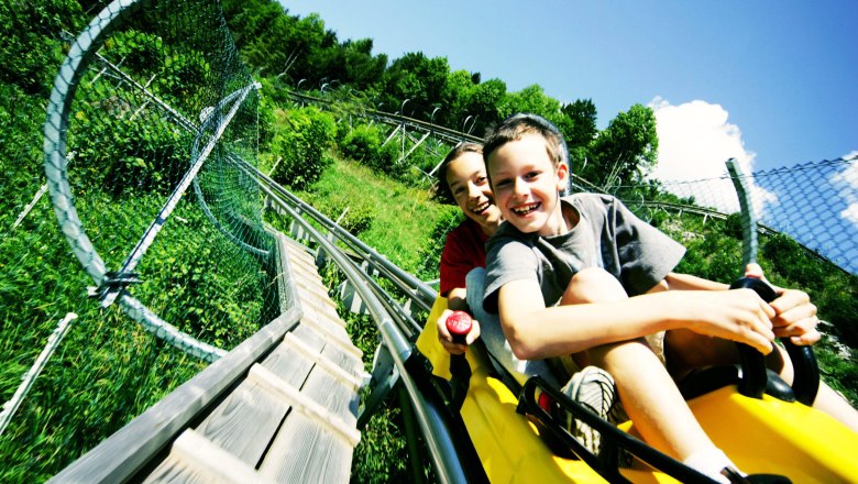 All-weather toboggan run Eibl-Jet in Türnitz, © weinfranz.at Two children laughing on a summer toboggan run through a green landscape.