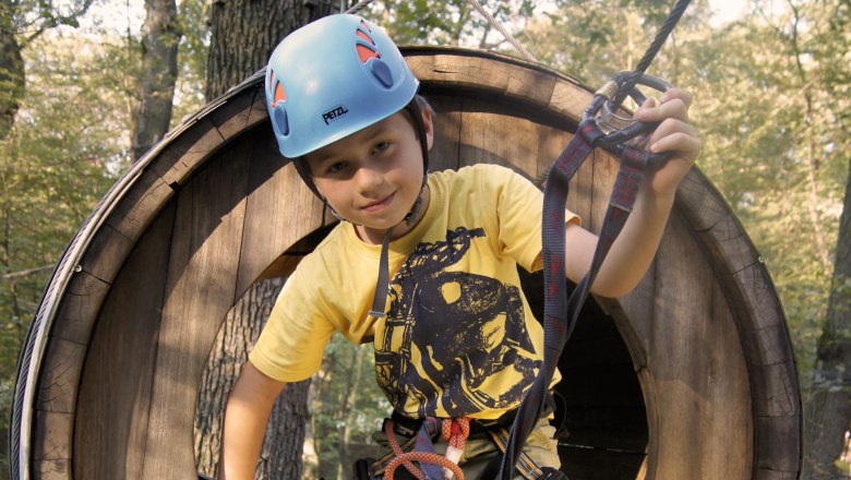 Climbing park, © Fam. Freudhofmaier A child with helmet and climbing equipment in a wooden climbing park tunnel.