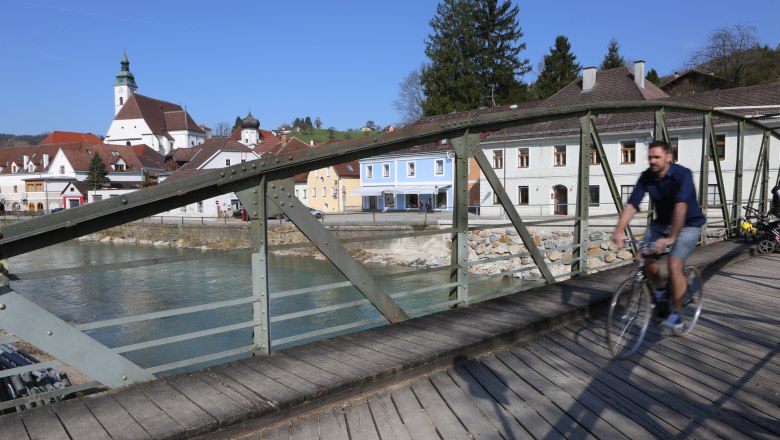 Töpper Bridge, © schwarz-koenig.at A cyclist crosses the Töpper Bridge in a picturesque town with a church and colorful houses in the background.
