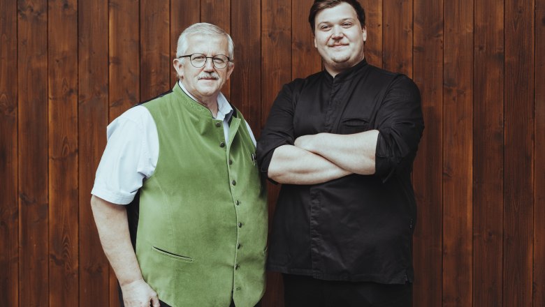 Reinhard Bürgmayr Junior and Senior, © Niederösterreich Werbung/David Schreiber Two men are standing in front of a wooden wall, one wearing a green vest, the other a black shirt.