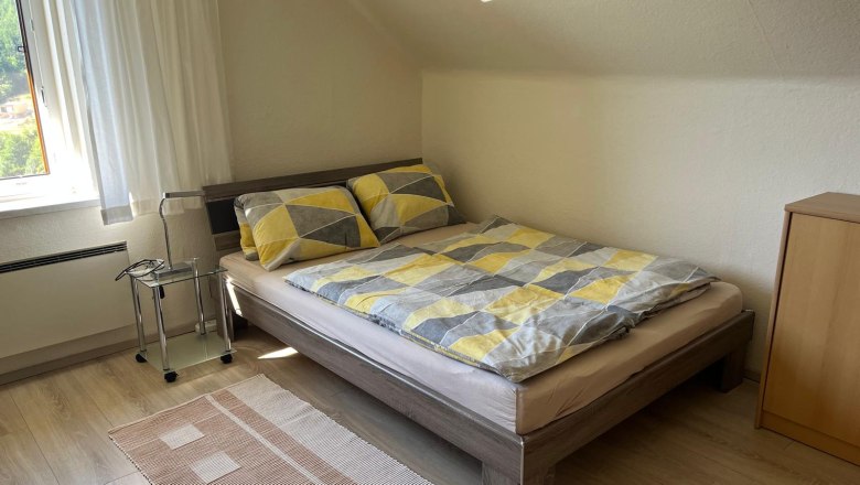 Apartments Riedl, © Familie Riedl A cozy bedroom with a double bed, patterned comforter covers, a small bedside table and a window with curtains.