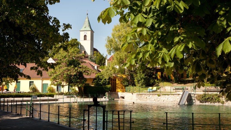 Thermal bath Bad Fischau-Brunn, © Marktgemeinde Bad Fischau-Brunn_Fotograf Lukas Dostal Bad Fischau-Brunn thermal baths with church in the background, surrounded by trees.